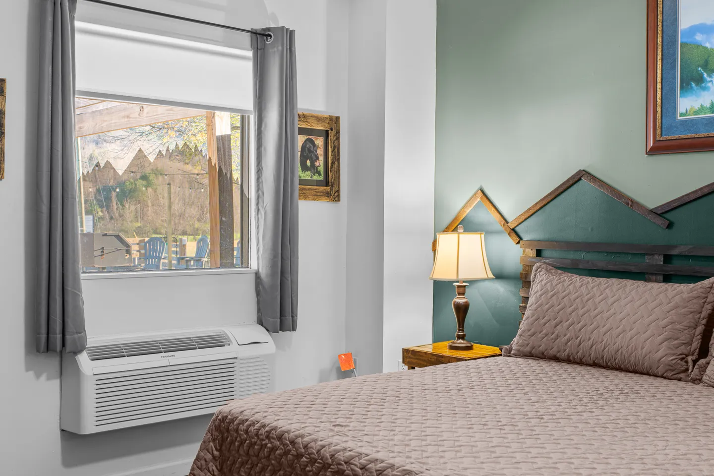 Bedroom featuring a made bed with a textured brown coverlet, a mountain-silhouette wooden headboard, a window with grey curtains, and an in-wall air conditioning unit.