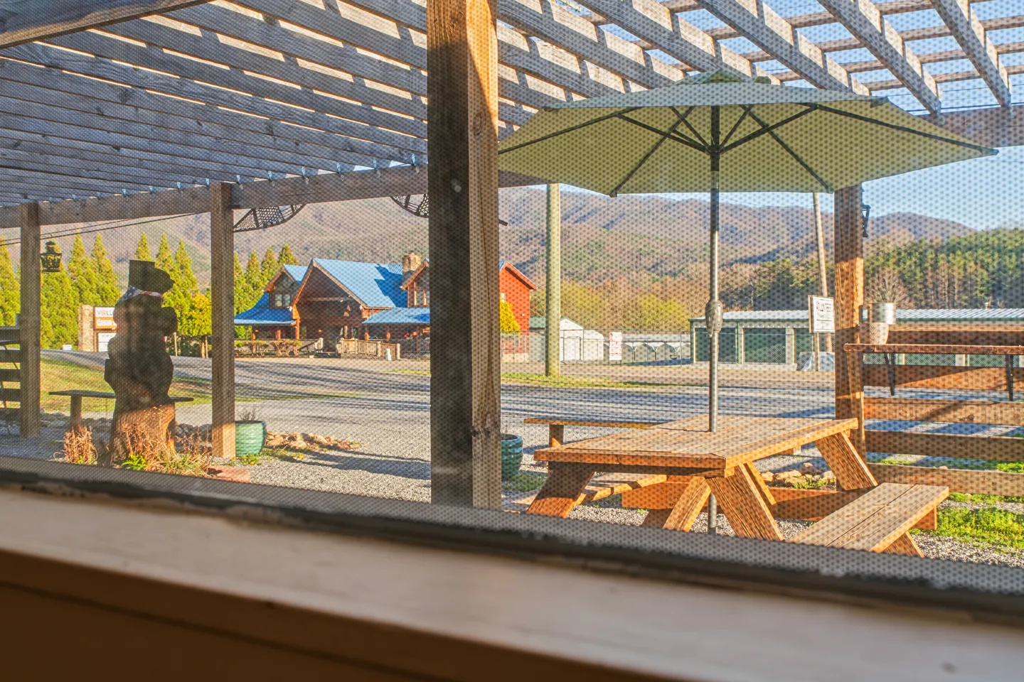 View through a window screen looking out at an outdoor wooden pergola with a picnic table and umbrella, with mountains and buildings in the background.