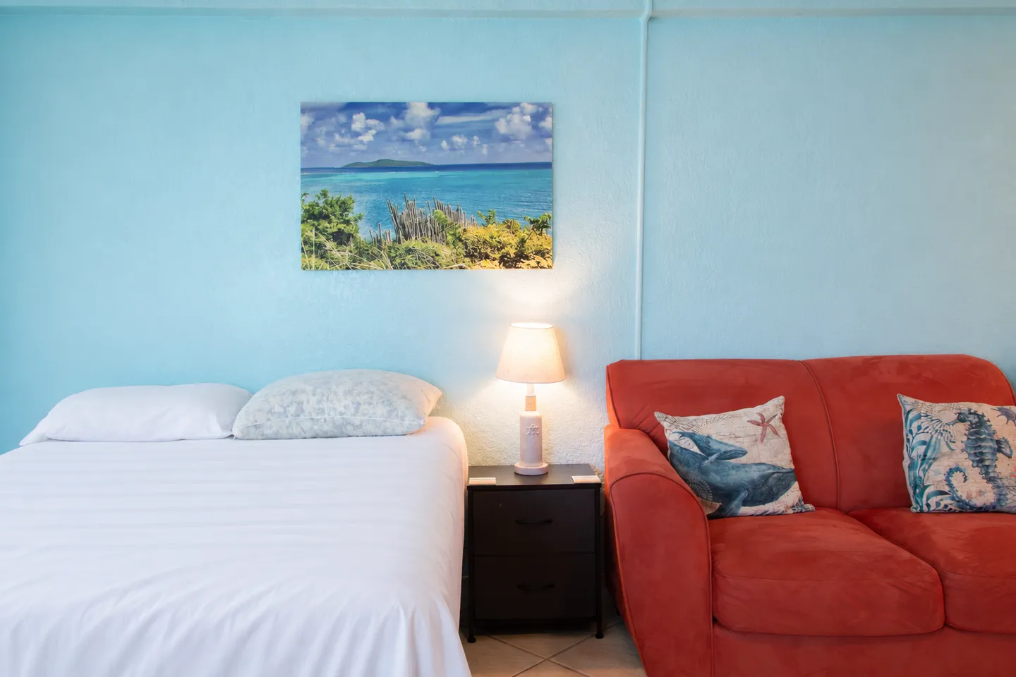 Bedroom area featuring a bed with white linens, a bedside table with a lamp, and an adjacent red sofa against a light blue wall.