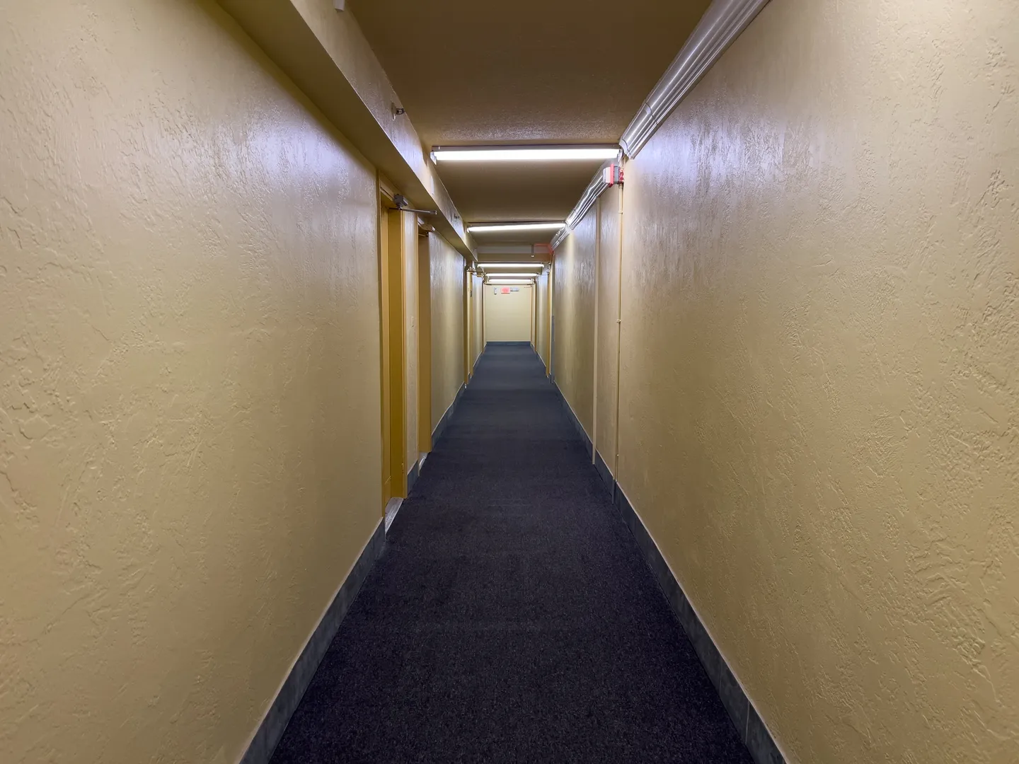 Straight-on view down a long interior hallway with yellow walls, dark carpeting, and fluorescent ceiling lights.