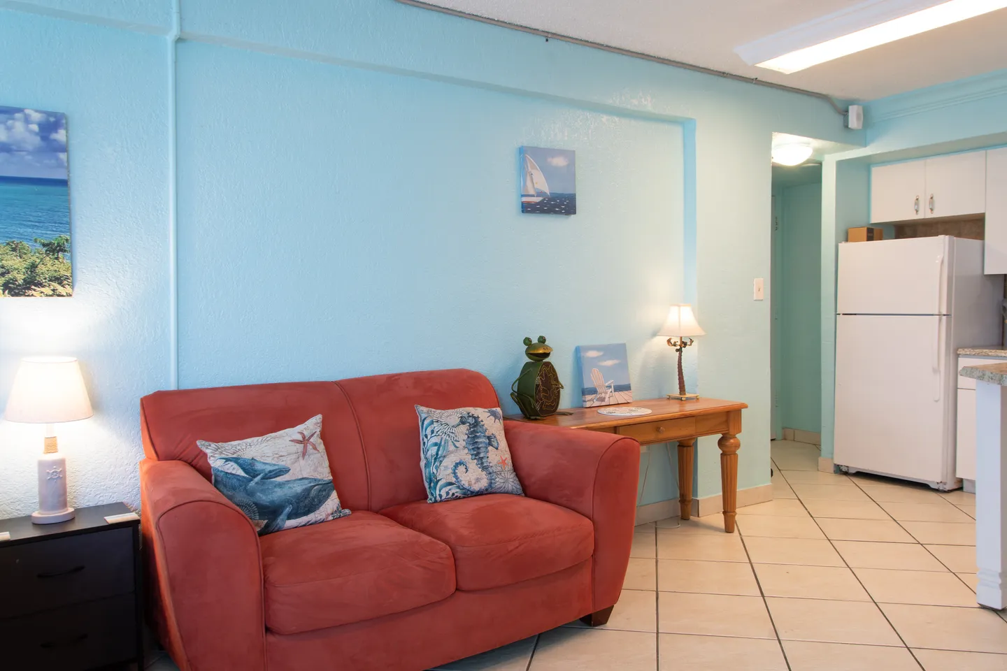 Living area with light blue walls, tiled floors, an orange-red loveseat, and an open view toward the kitchen.