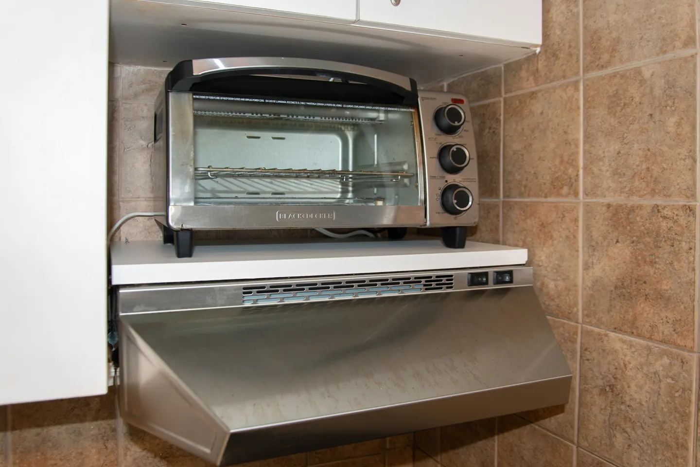 A Black and Decker toaster oven sitting on a shelf above a stainless steel range hood against brown tiled walls.