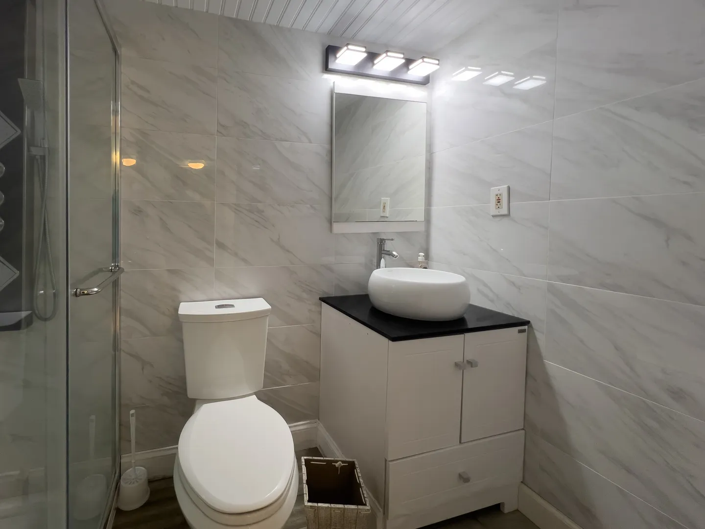 Bathroom with marble-patterned wall tiles, a toilet, vanity with vessel sink, and glass shower door.