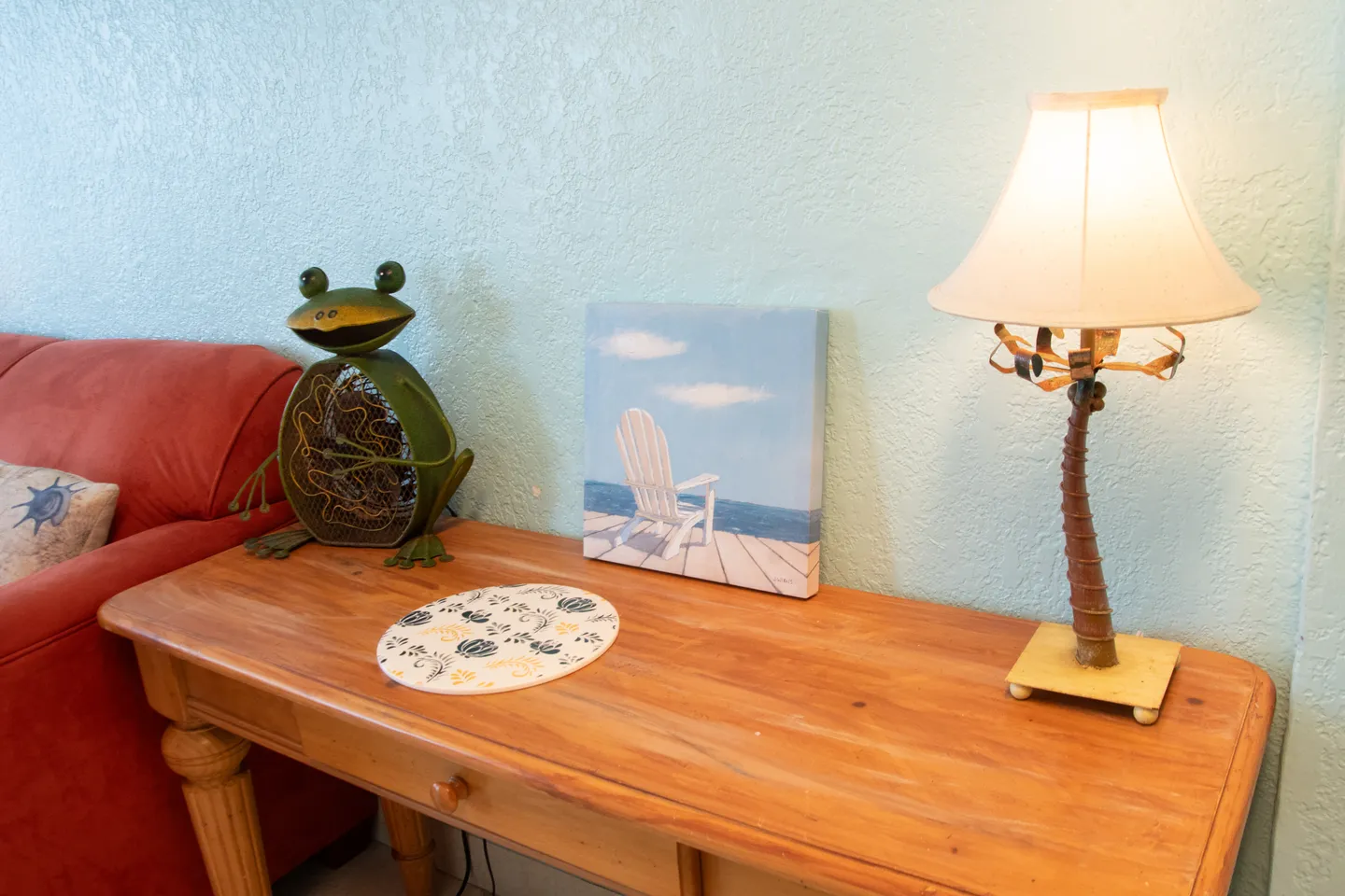 Wooden side table featuring a palm tree lamp, a small beach canvas, a frog-shaped decorative fan, and a coaster next to a red sofa.