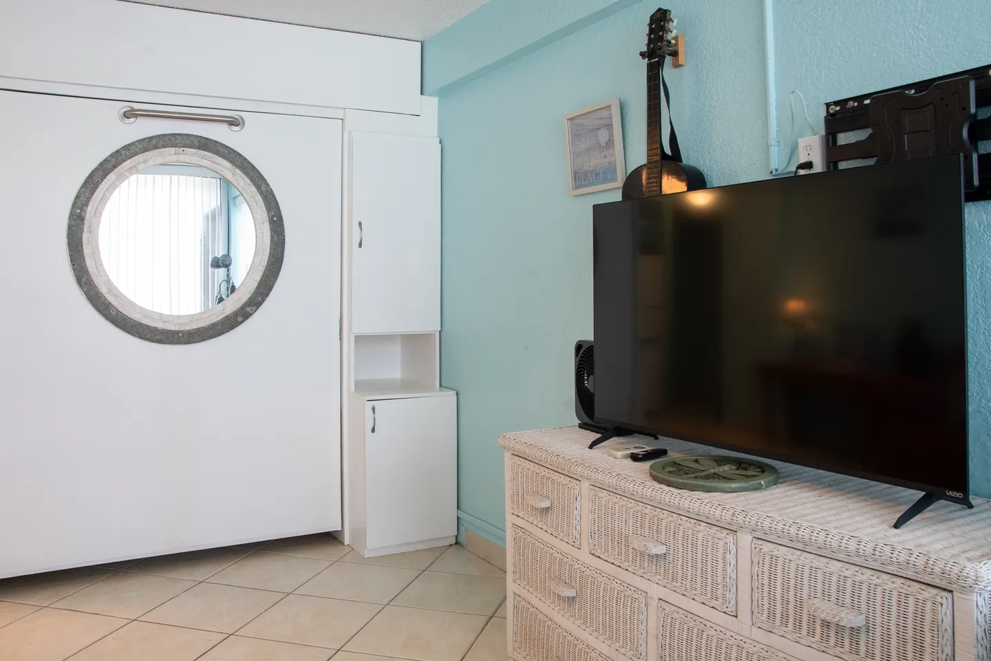 Living space featuring a closed white Murphy bed with a porthole mirror, tiled flooring, and a flat-screen television on a white wicker dresser against a light blue wall.
