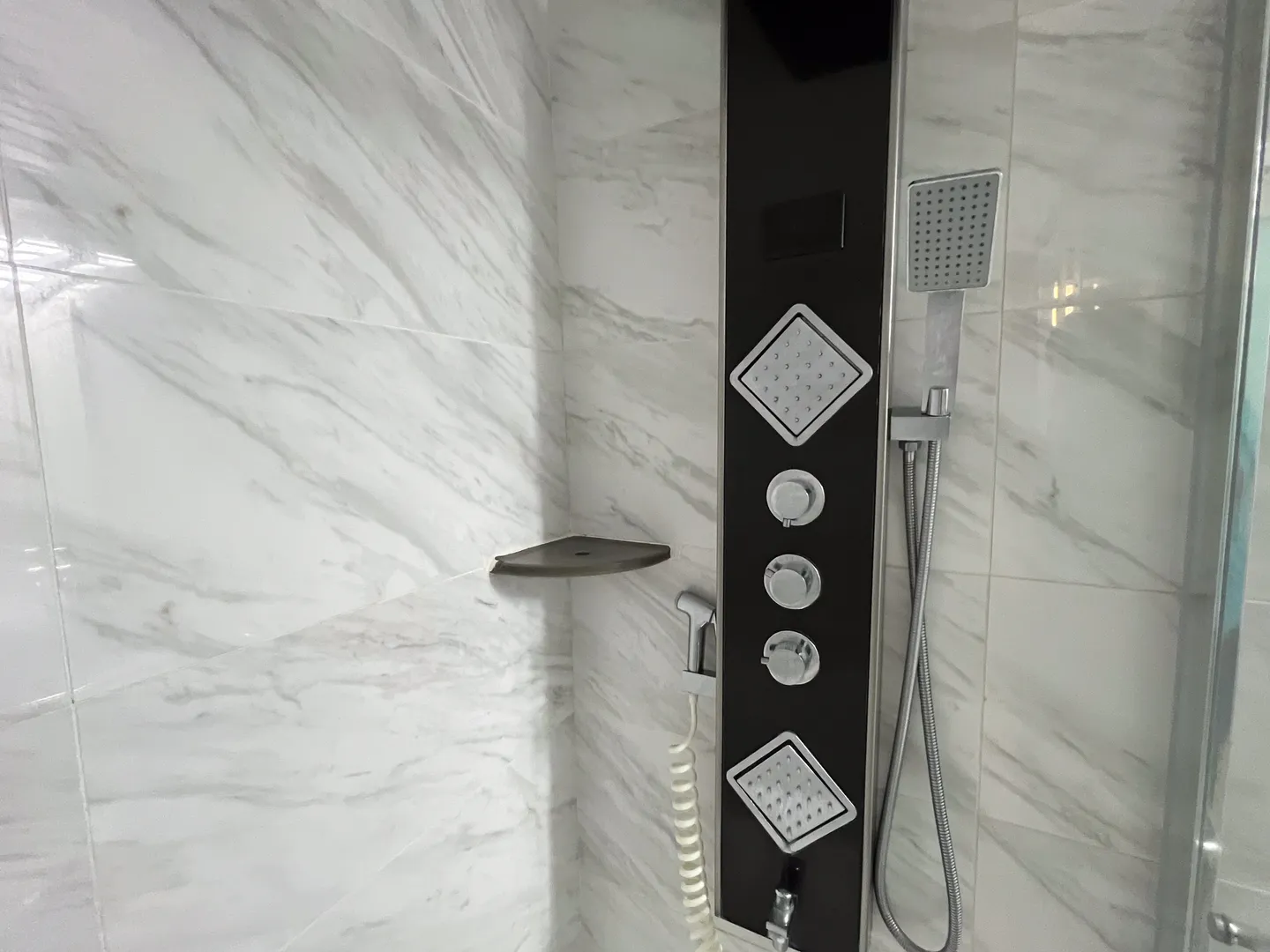 Close-up of a shower with marble-look wall tiles and a modern black shower panel featuring body jets and a handheld sprayer.