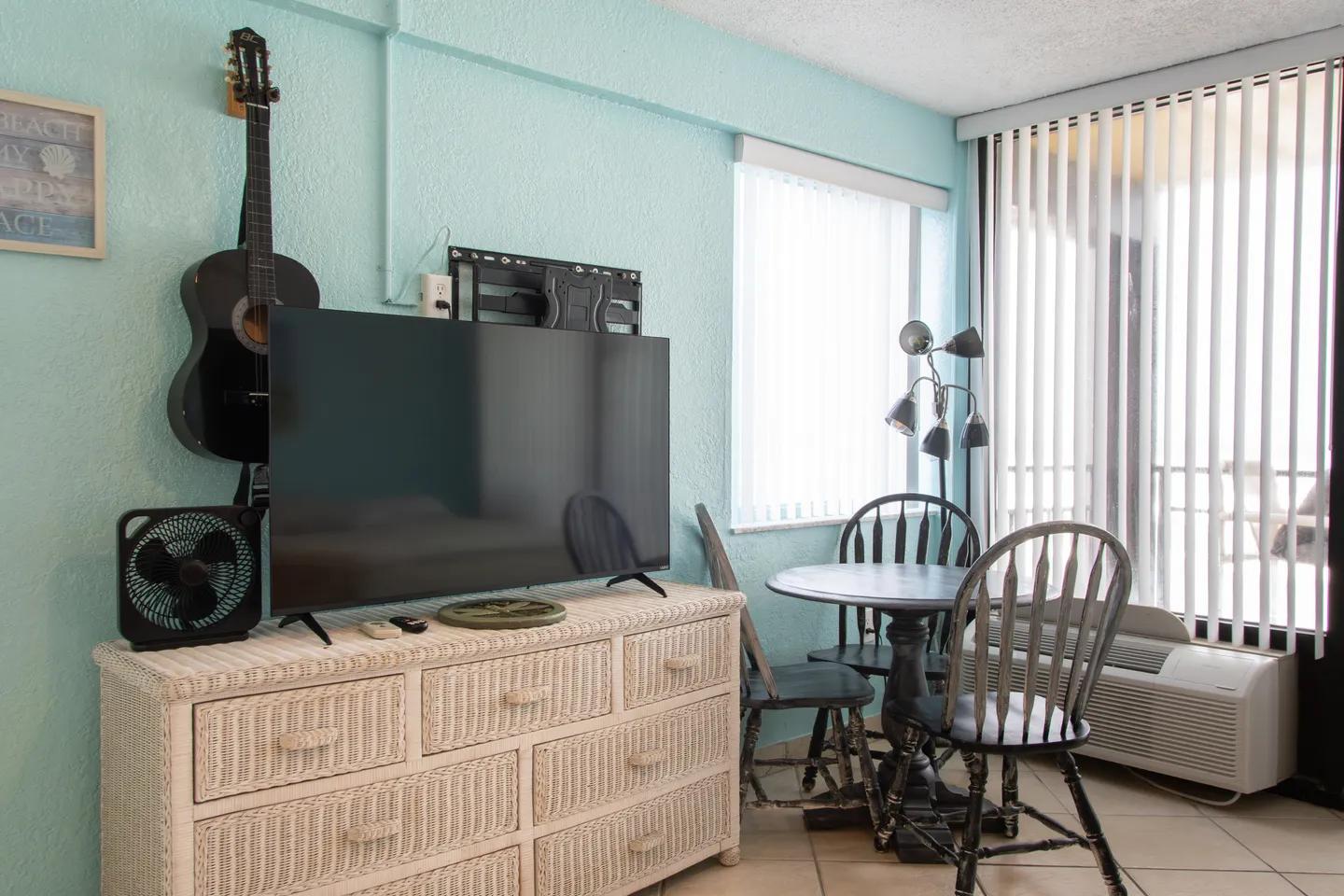 Living area featuring a flat-screen TV on a white wicker dresser beside a small dining table and chairs with sliding doors in the background.