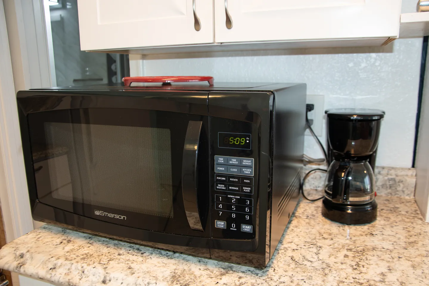 A black Emerson countertop microwave and a small black drip coffee maker sitting on a speckled granite-style countertop beneath white kitchen cabinets.