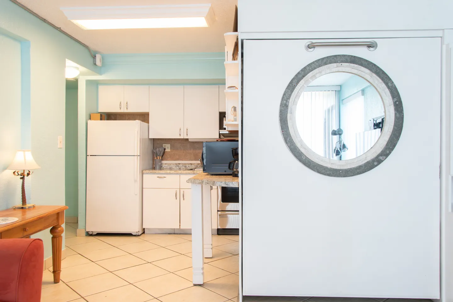 Living area looking into a kitchen with light blue walls, white cabinets, white refrigerator, and tiled flooring.