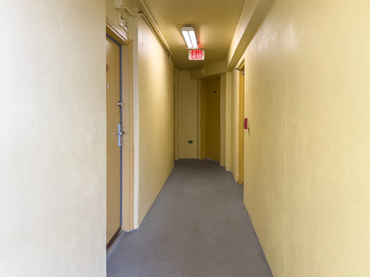 Indoor hallway with pale yellow walls, a light gray floor, several entry doors, and a red illuminated exit sign overhead.