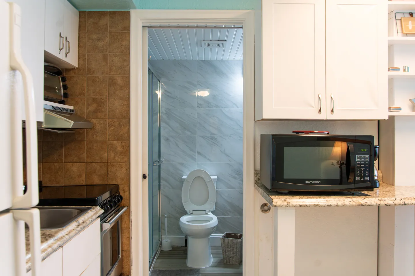 A doorway from a kitchen area with cabinets and appliances opening into a tiled bathroom featuring a toilet and glass shower door.