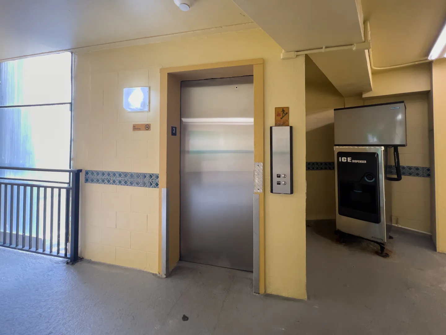 Elevator landing with stainless steel door and an ice dispenser alcove on a yellow cinderblock hallway.