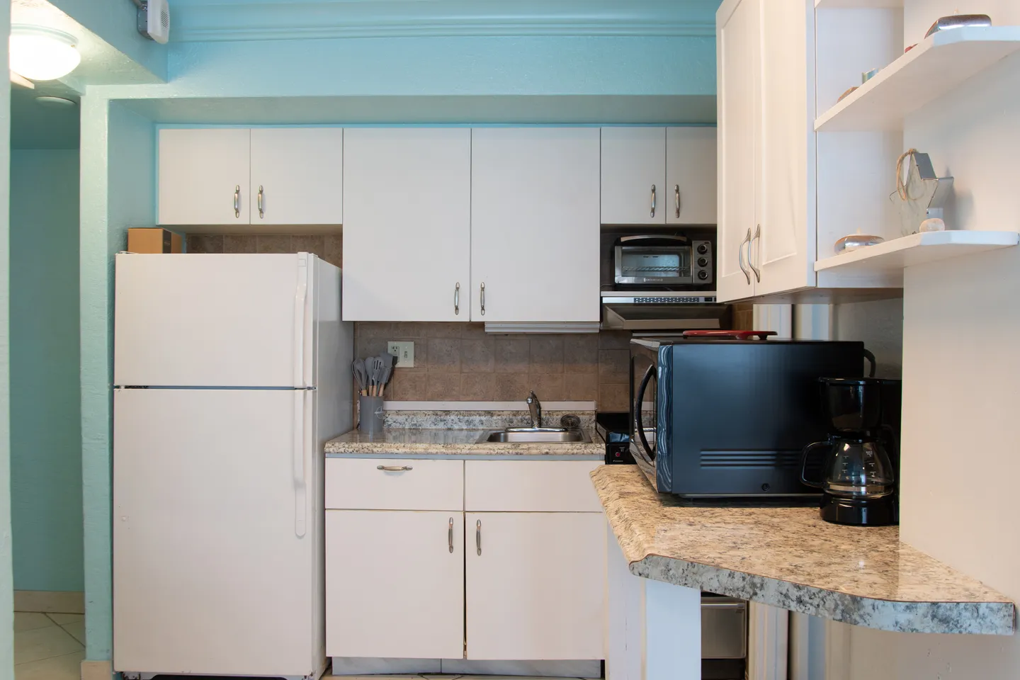 Kitchenette with white cabinets, speckled countertops, a full-size white refrigerator, microwave, and small sink against light blue walls.