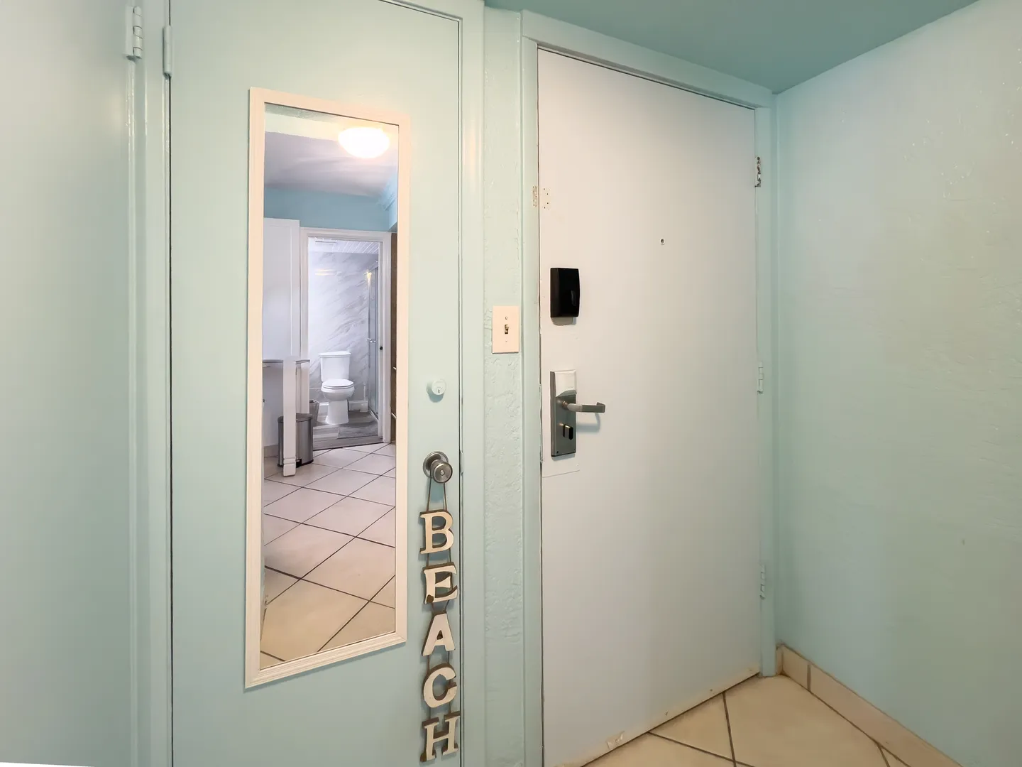 Entryway with light blue walls, a white front door featuring an electronic lock, tile flooring, and a full-length mirror reflecting a bathroom.