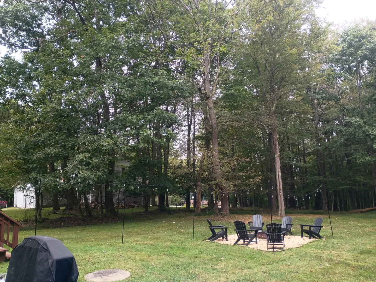 A backyard lawn bordered by dense woods with a gravel fire pit area surrounded by chairs and a covered grill nearby.
