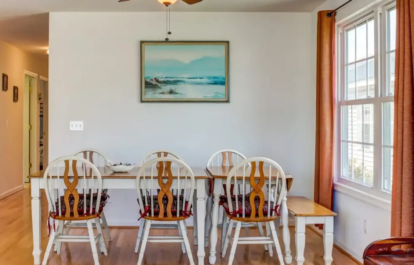 Dining area with a wooden table, white and wood chairs with red cushions, a painting on the wall, and a window with orange curtains.