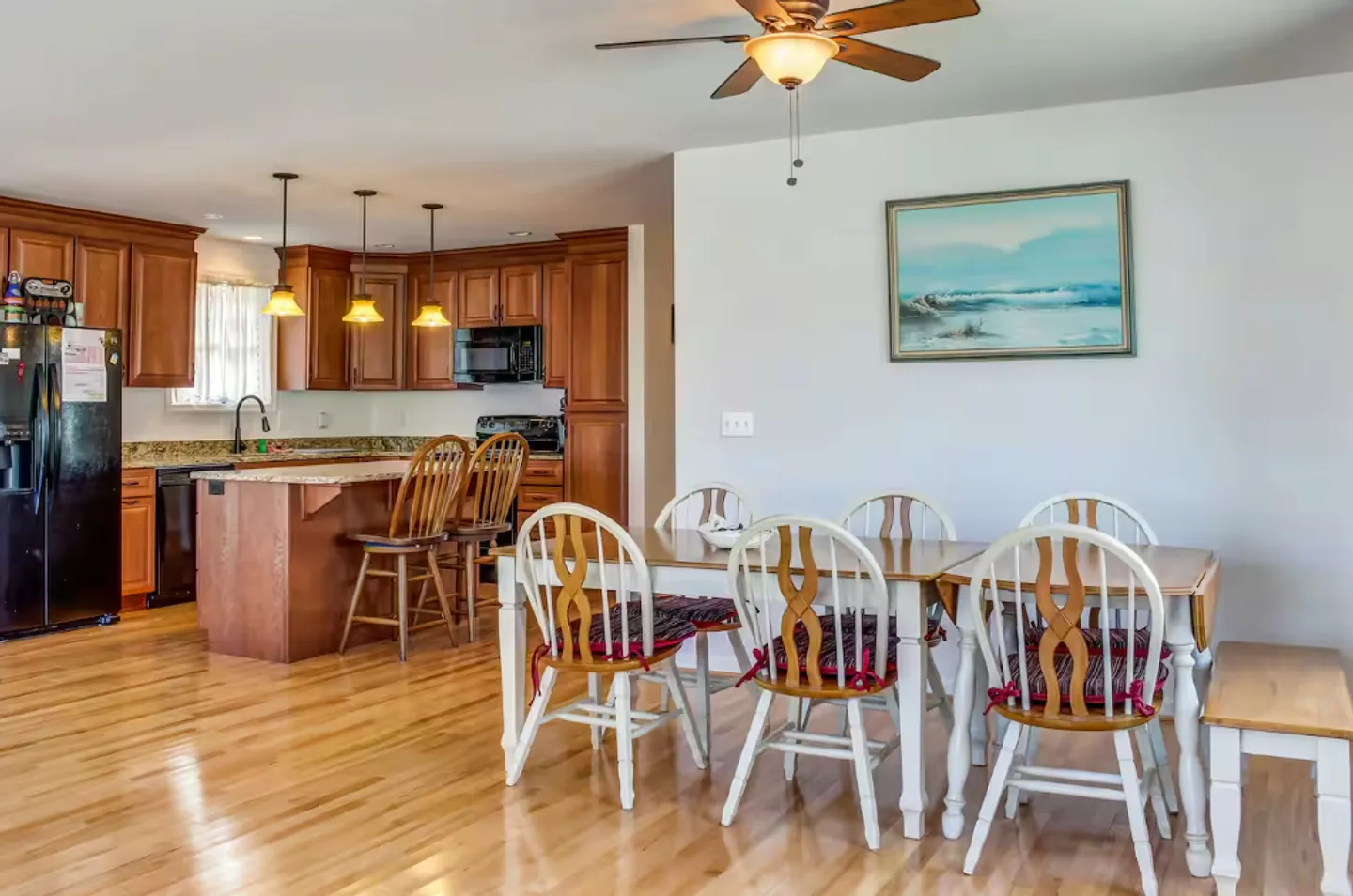 Dining area with a wooden table and white-framed chairs, open to a kitchen with wood cabinets.