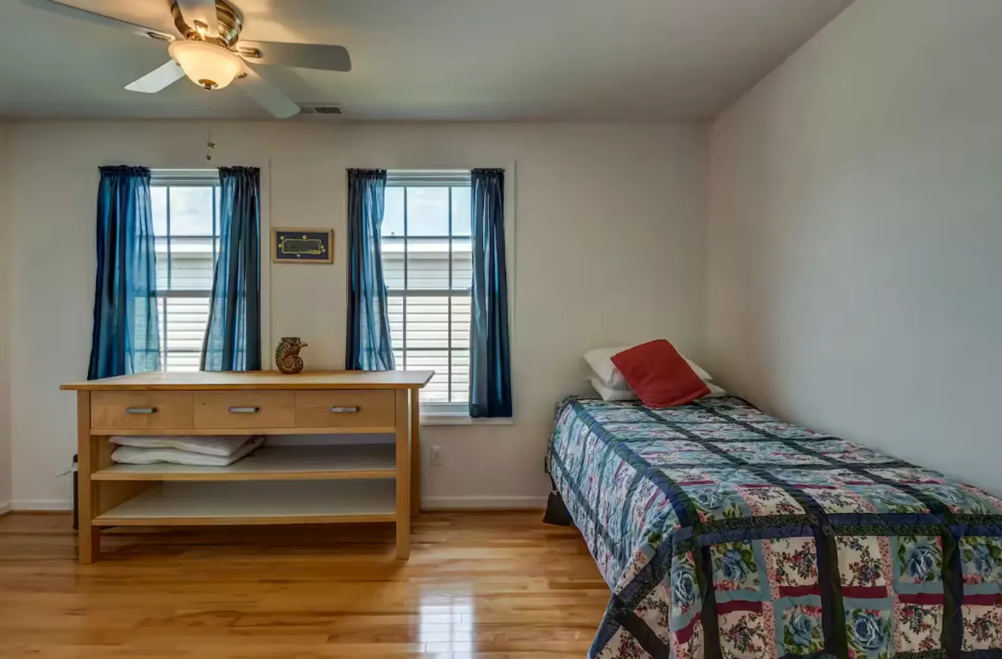 Bedroom with hardwood floors, a single bed covered in a patterned quilt, and a light wood sideboard beneath two windows with blue curtains.