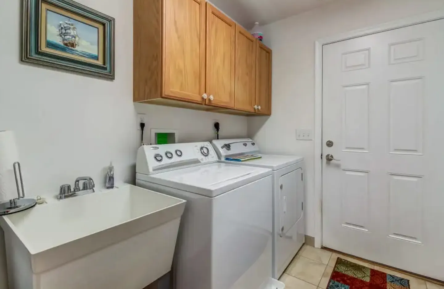 Laundry room with a utility sink, white washer and dryer, and upper wooden cabinets beside a white interior door.