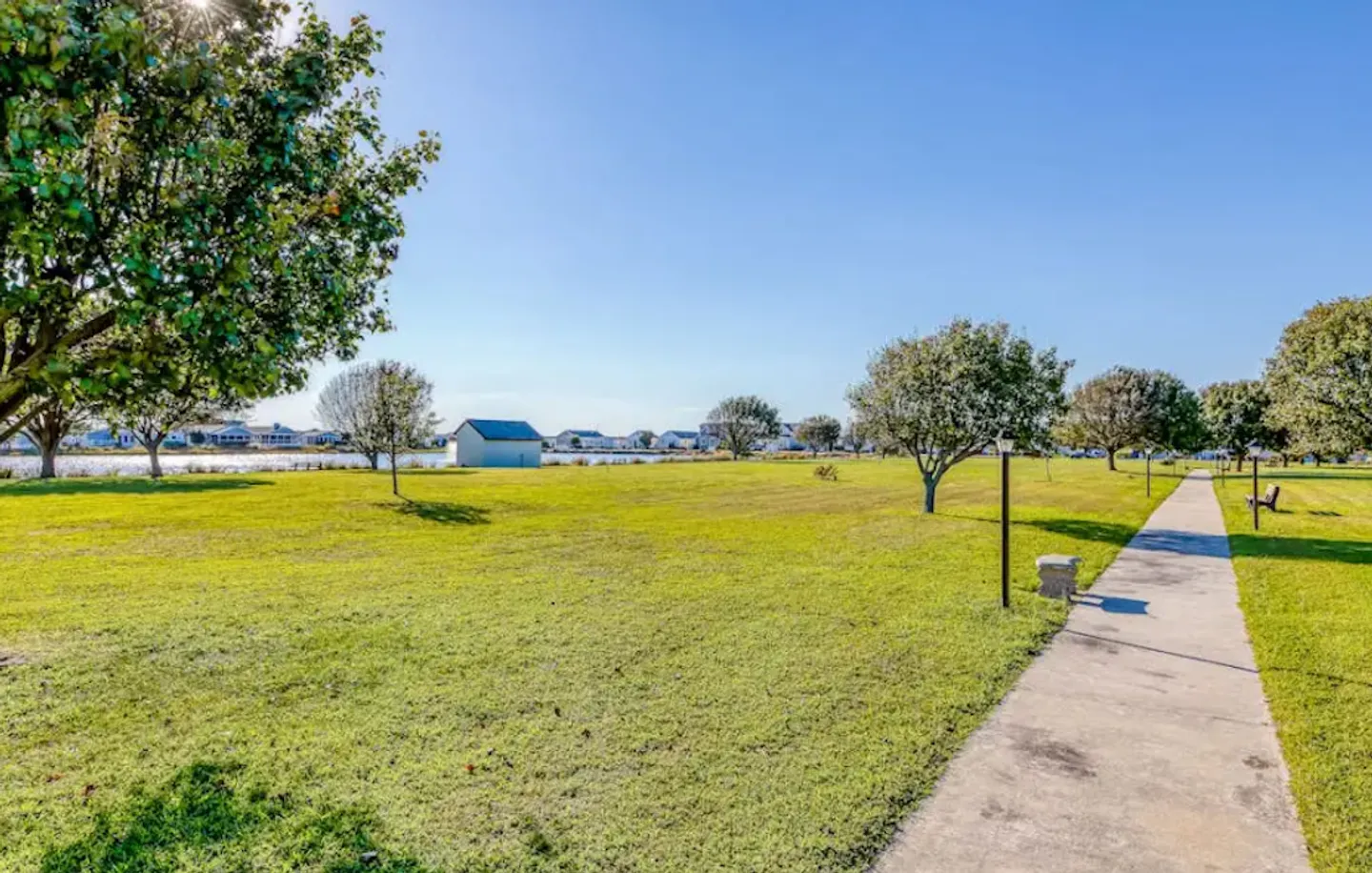 A concrete walkway cutting through a large grassy lawn toward a pond or lake under a clear blue sky.