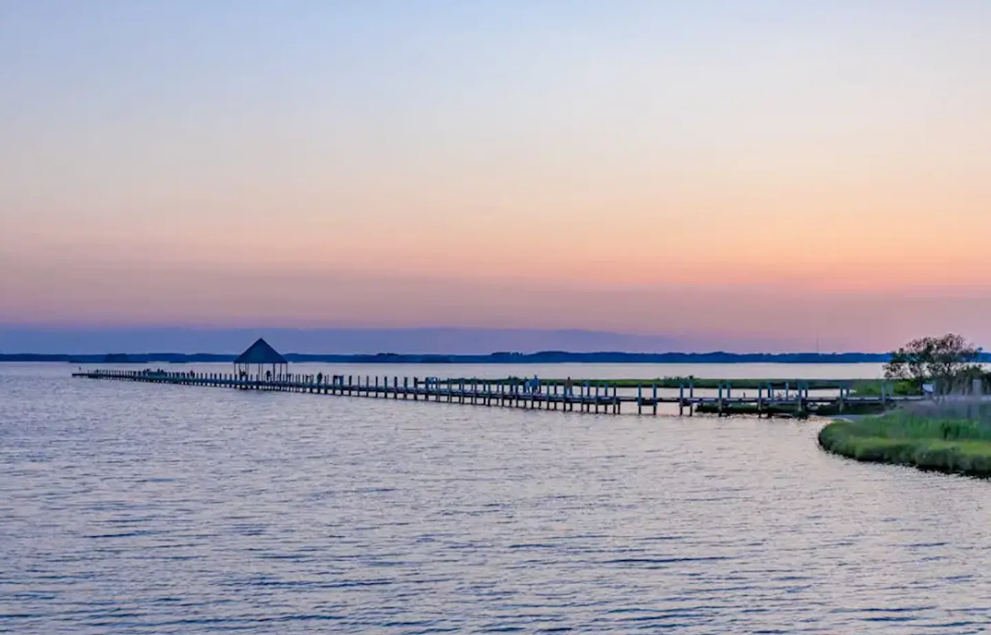 A long wooden pier with a covered gazebo extending into calm water during sunset.