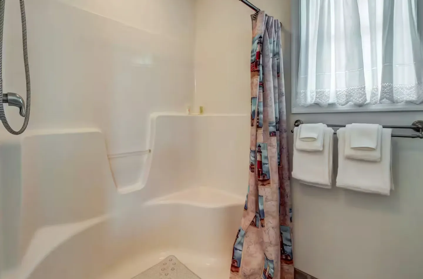 Bathroom with a white molded shower stall featuring a built-in corner seat, a patterned shower curtain, and a towel rack with folded white towels next to a curtained window.