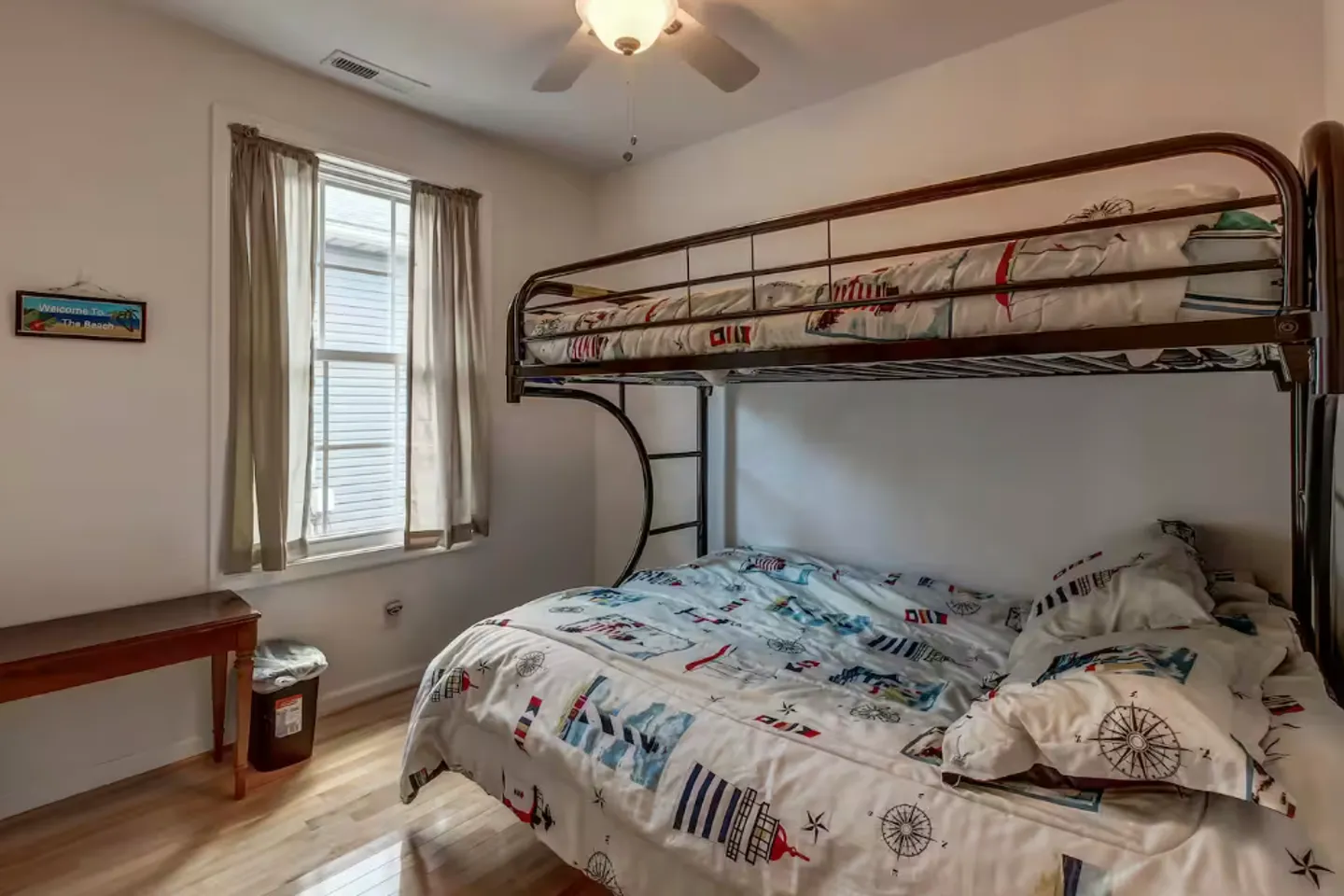 Bedroom with wooden floors and metal bunk beds featuring nautical bedding next to a curtained window.
