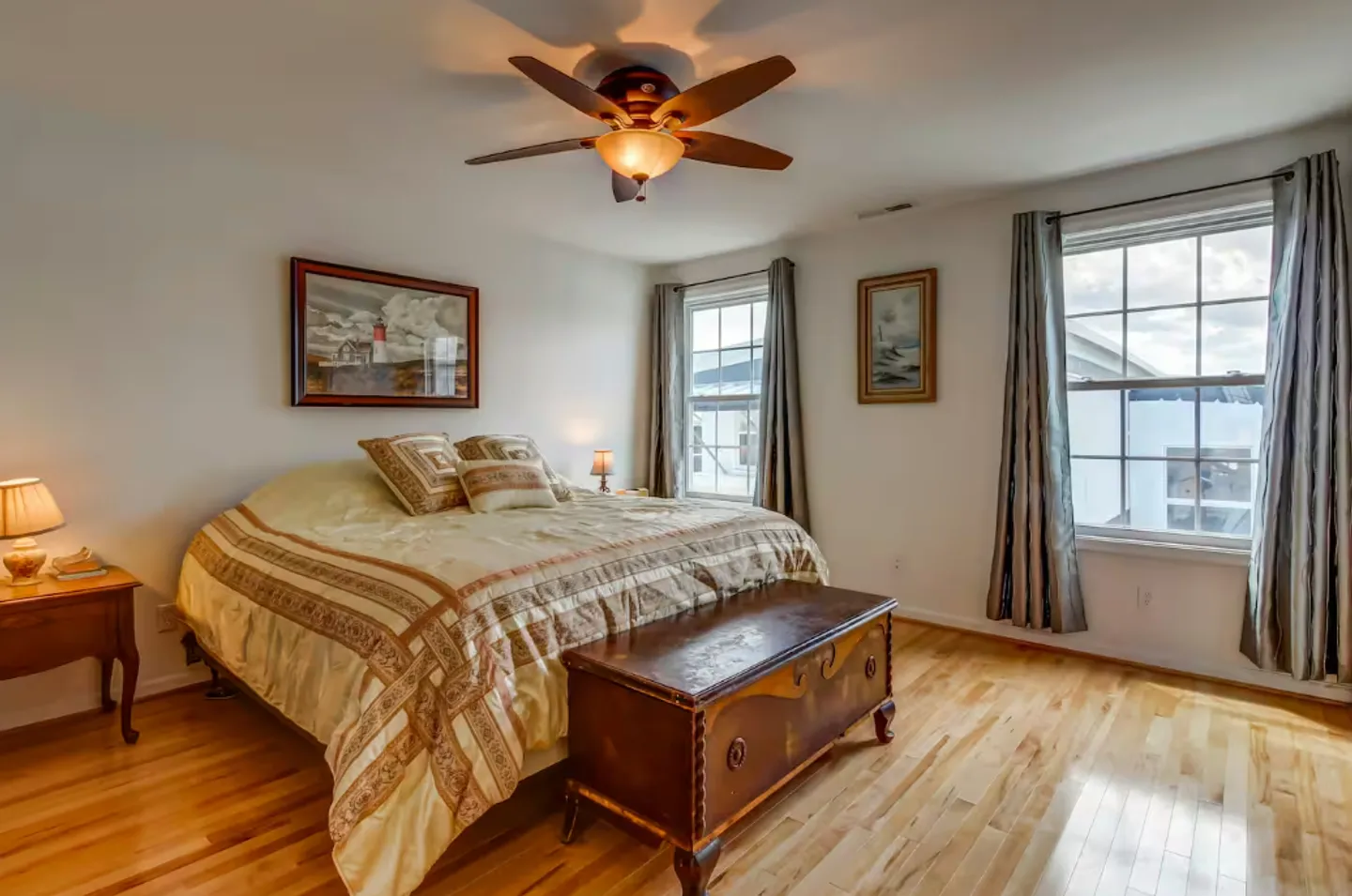 Bedroom with hardwood floors, a large bed with neutral bedding, a decorative trunk at the foot, and two windows with curtains.