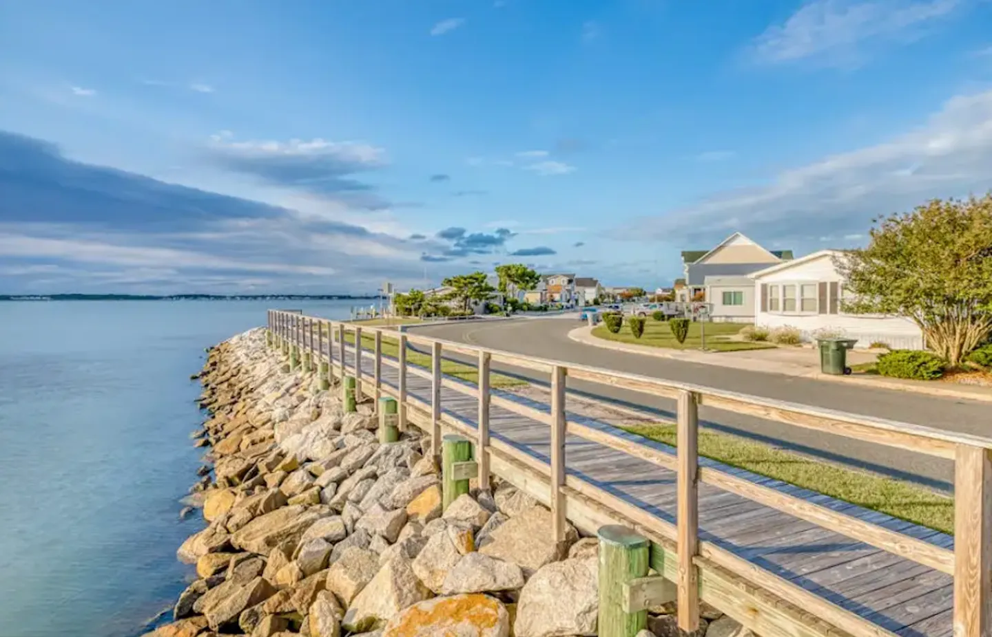 A wooden boardwalk with railings runs along a rocky shoreline next to a paved coastal street and houses.