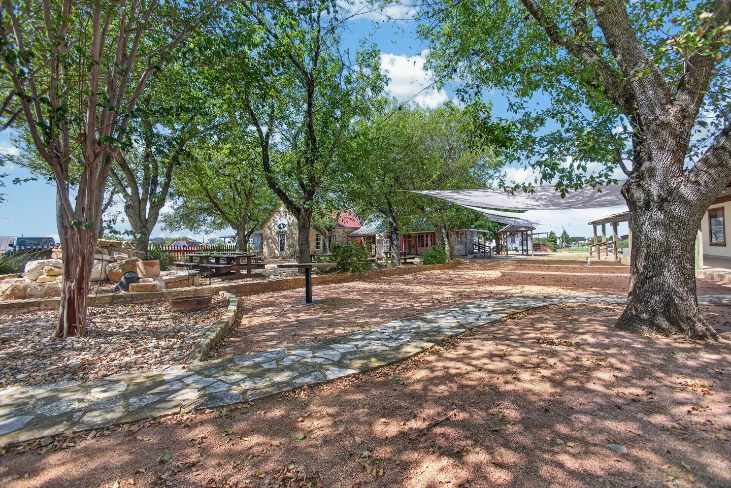 A wide gravel courtyard with stone walkways, shade trees, picnic tables, and rustic buildings under a partly cloudy sky.