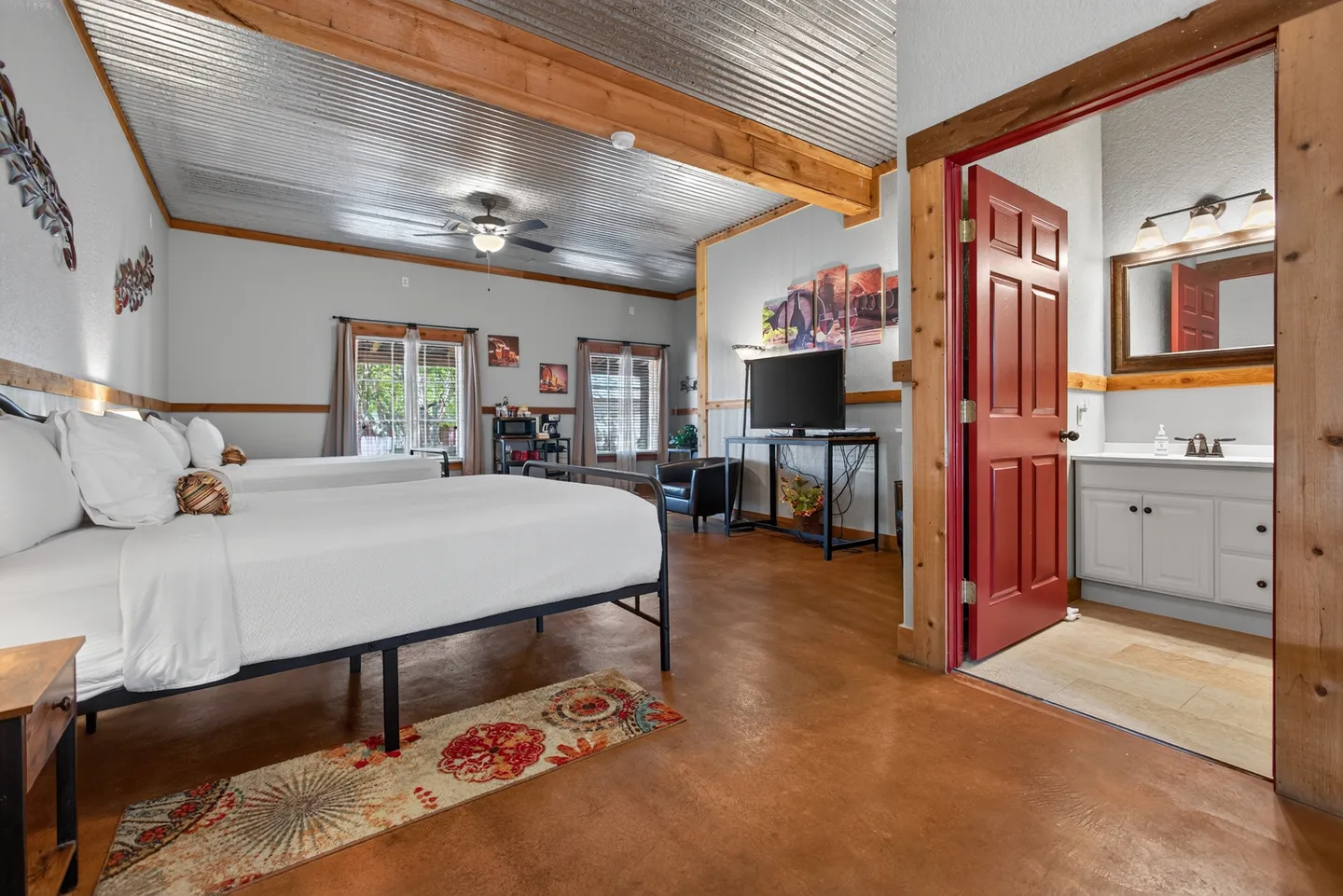 Bedroom with two beds, corrugated metal ceiling with wooden beams, stained concrete floors, and an open doorway leading to an ensuite bathroom.