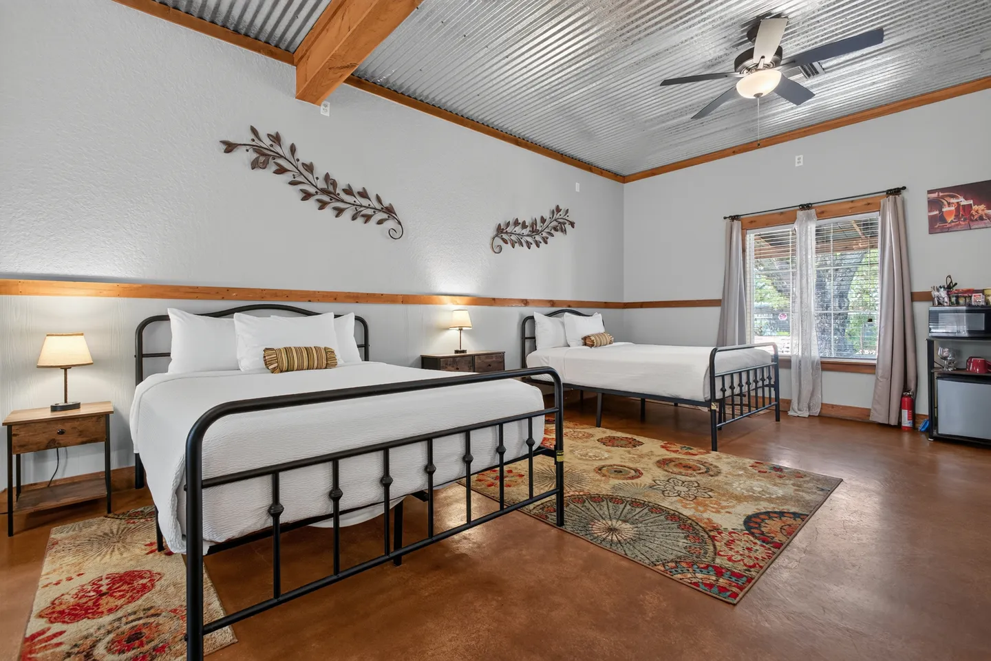 Spacious bedroom featuring two wrought-iron frame beds, a corrugated metal ceiling with wooden beams, patterned area rugs, and smooth concrete floors.