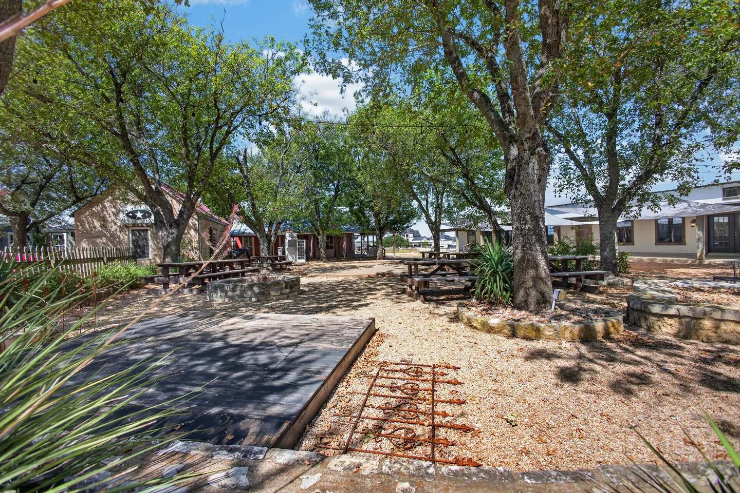 Outdoor courtyard with gravel ground, large shade trees, picnic tables, and a small wooden platform.