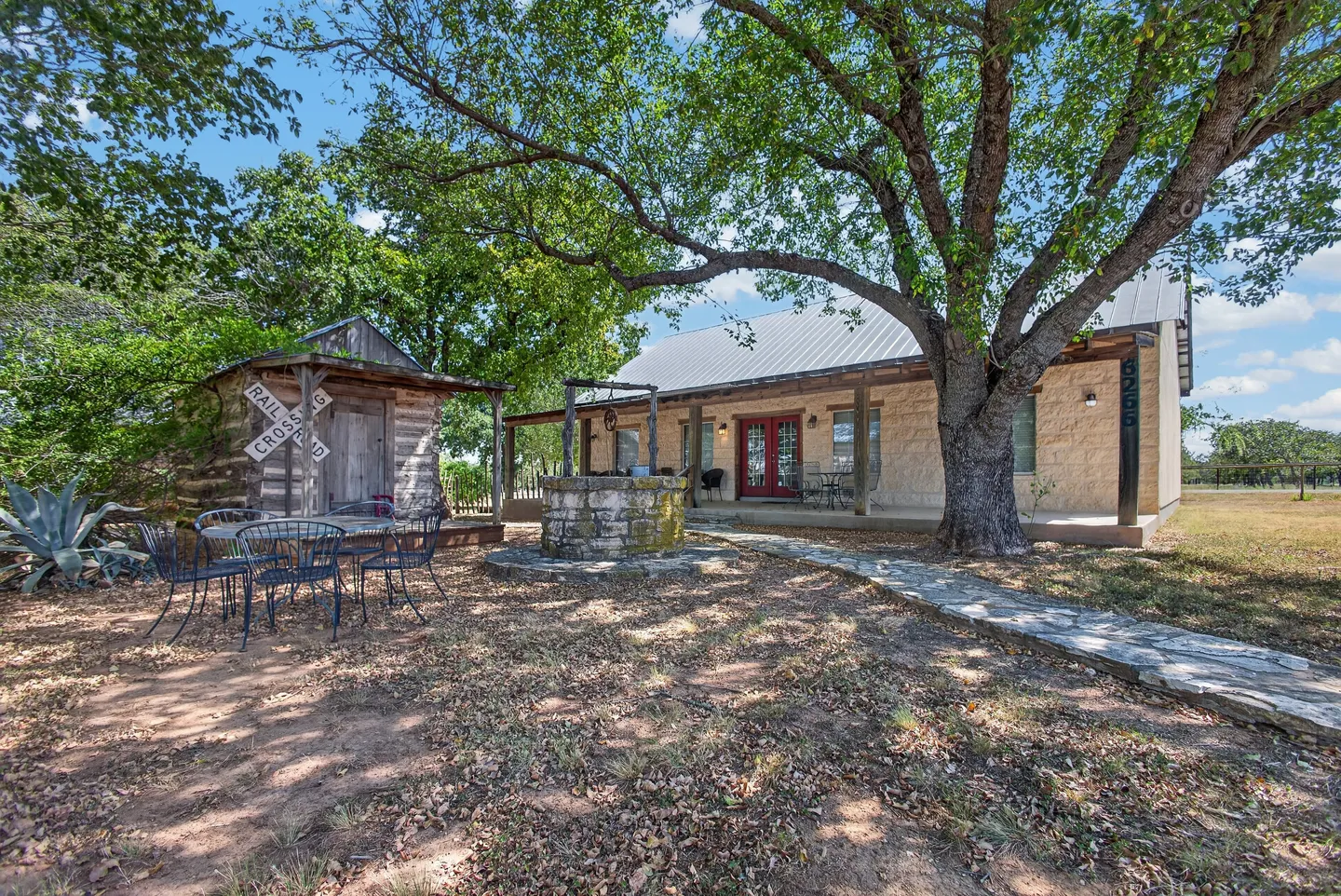 Exterior of a stone cottage with a covered front porch, stone wishing well, small rustic shed, and an outdoor patio dining set under a large shade tree.
