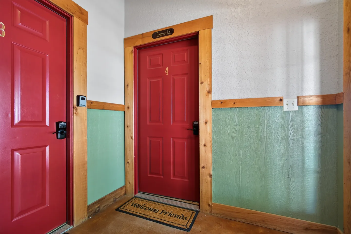 A hallway featuring red doors with wood trim and keyless locks, paneled green wainscoting, and a doormat that reads Welcome Friends.