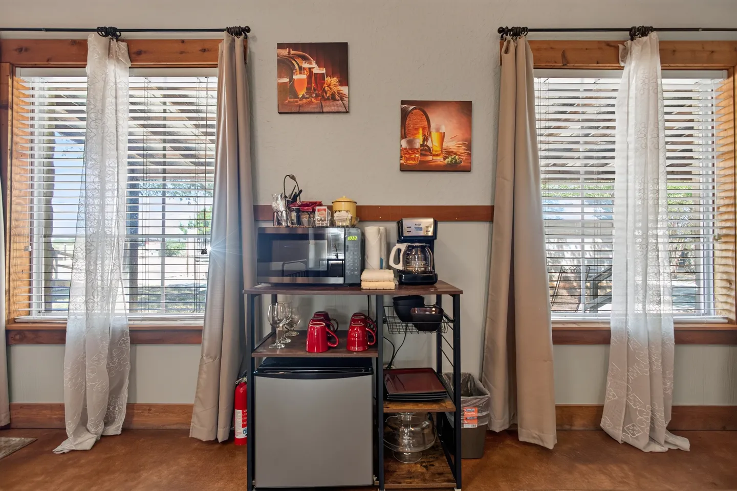 A beverage and snack station featuring a mini fridge, microwave, and coffee maker flanked by two windows with wooden trim.