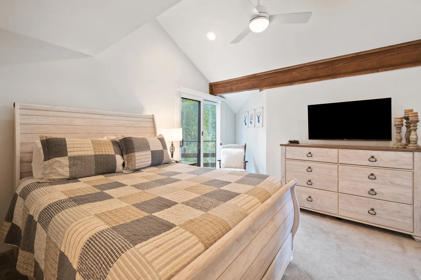 Bedroom with vaulted ceiling, exposed wooden beam, made bed with patterned quilt, white dresser, and mounted TV.