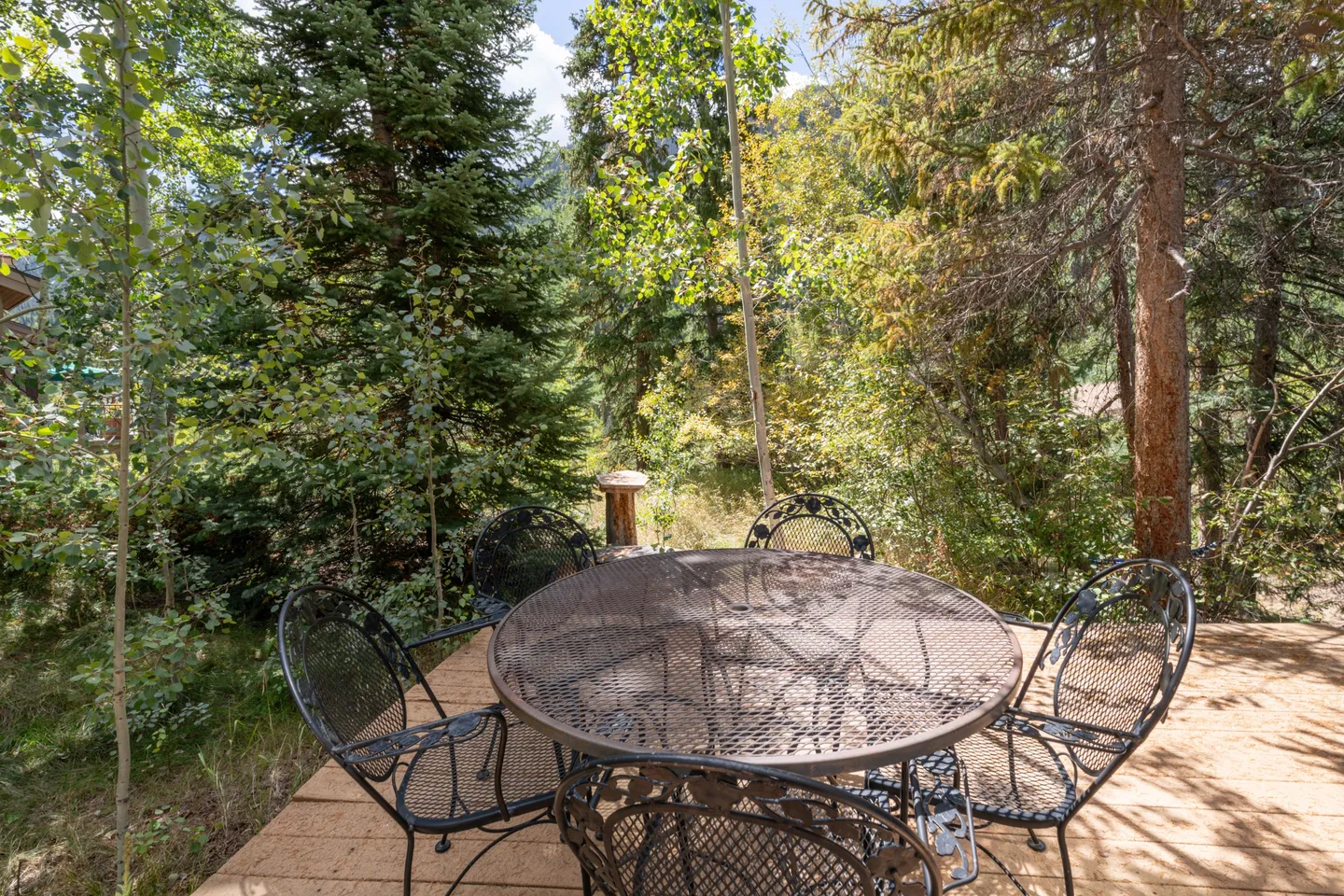 Outdoor wooden deck featuring a round metal mesh patio table and matching chairs, surrounded by dense pine and aspen trees under bright daylight.