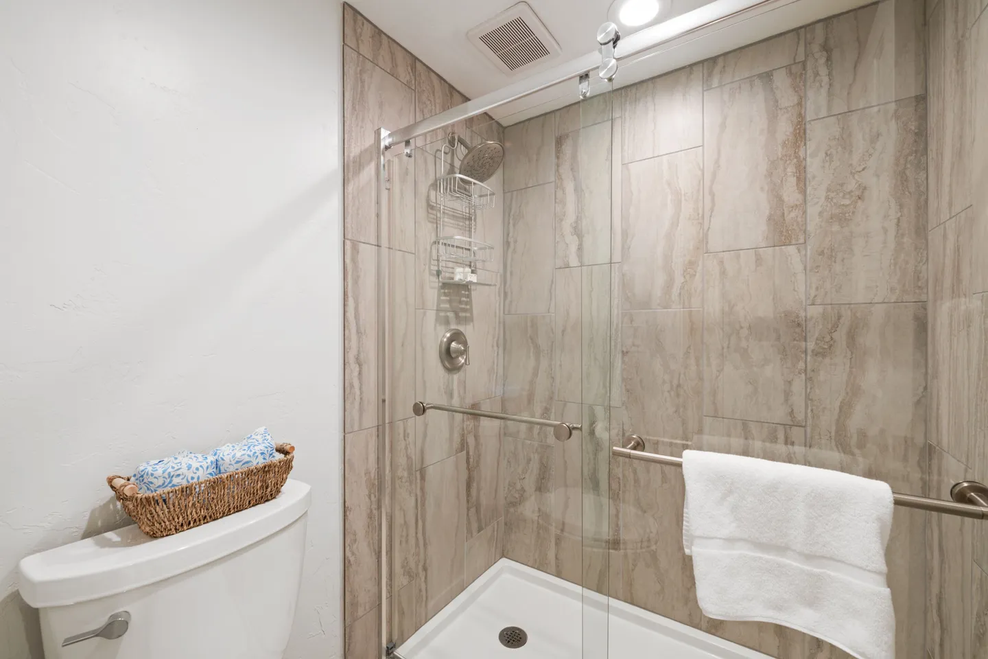 Bathroom with a glass-enclosed walk-in shower featuring large neutral tiles and a partial view of the toilet tank with a decorative basket.
