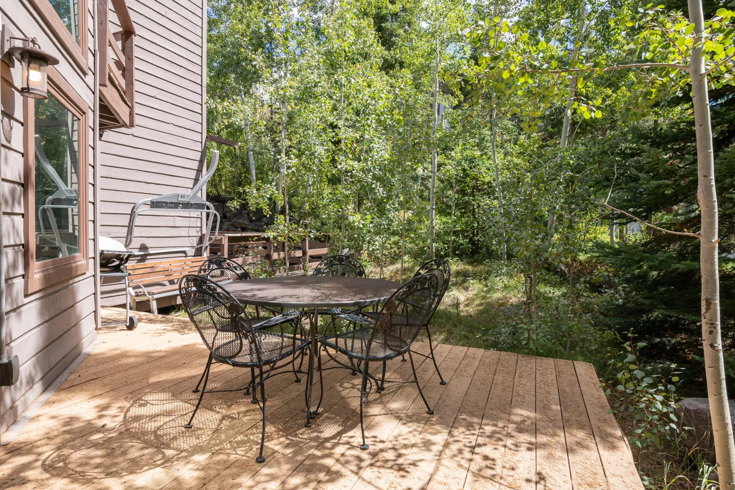 Outdoor wood deck with a round patio table, chairs, a barbecue grill, and a ski lift chair bench surrounded by leafy trees.