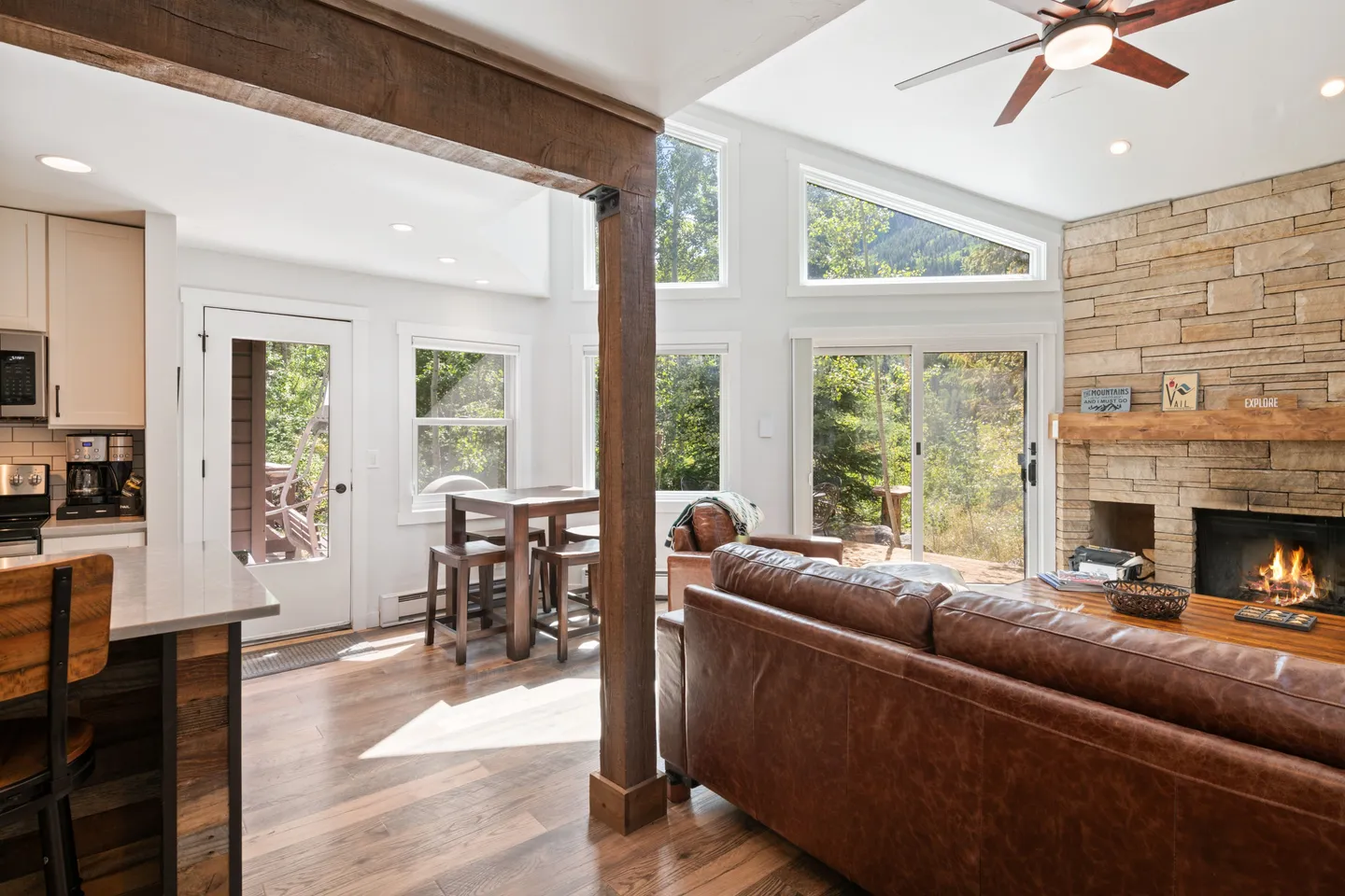 Open living area with a brown leather couch, stone fireplace, wood dining set, and kitchen counter under vaulted ceilings.