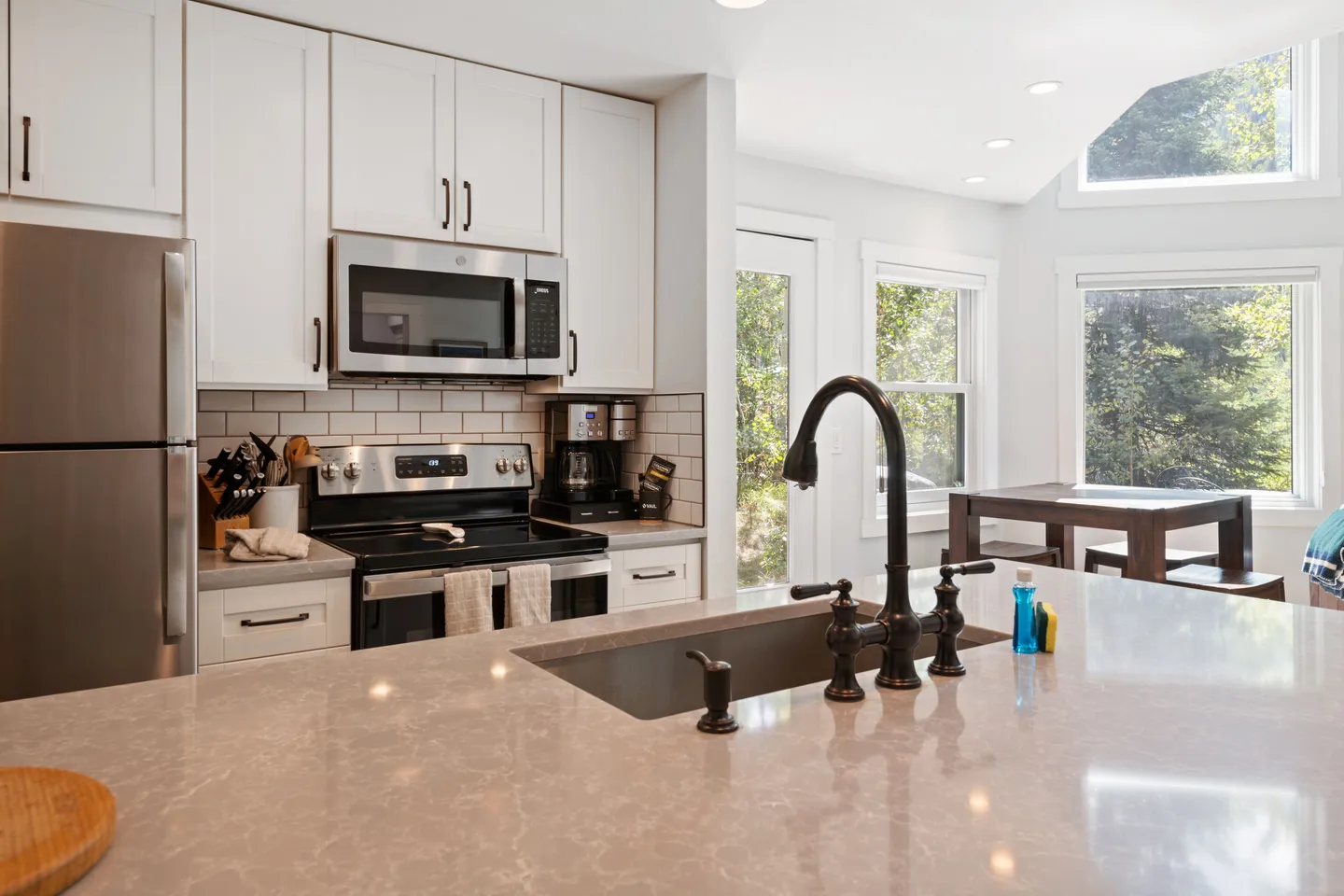 A kitchen with white shaker cabinets, stainless steel appliances, a center island with an undermount sink, and an adjacent dining nook with windows.