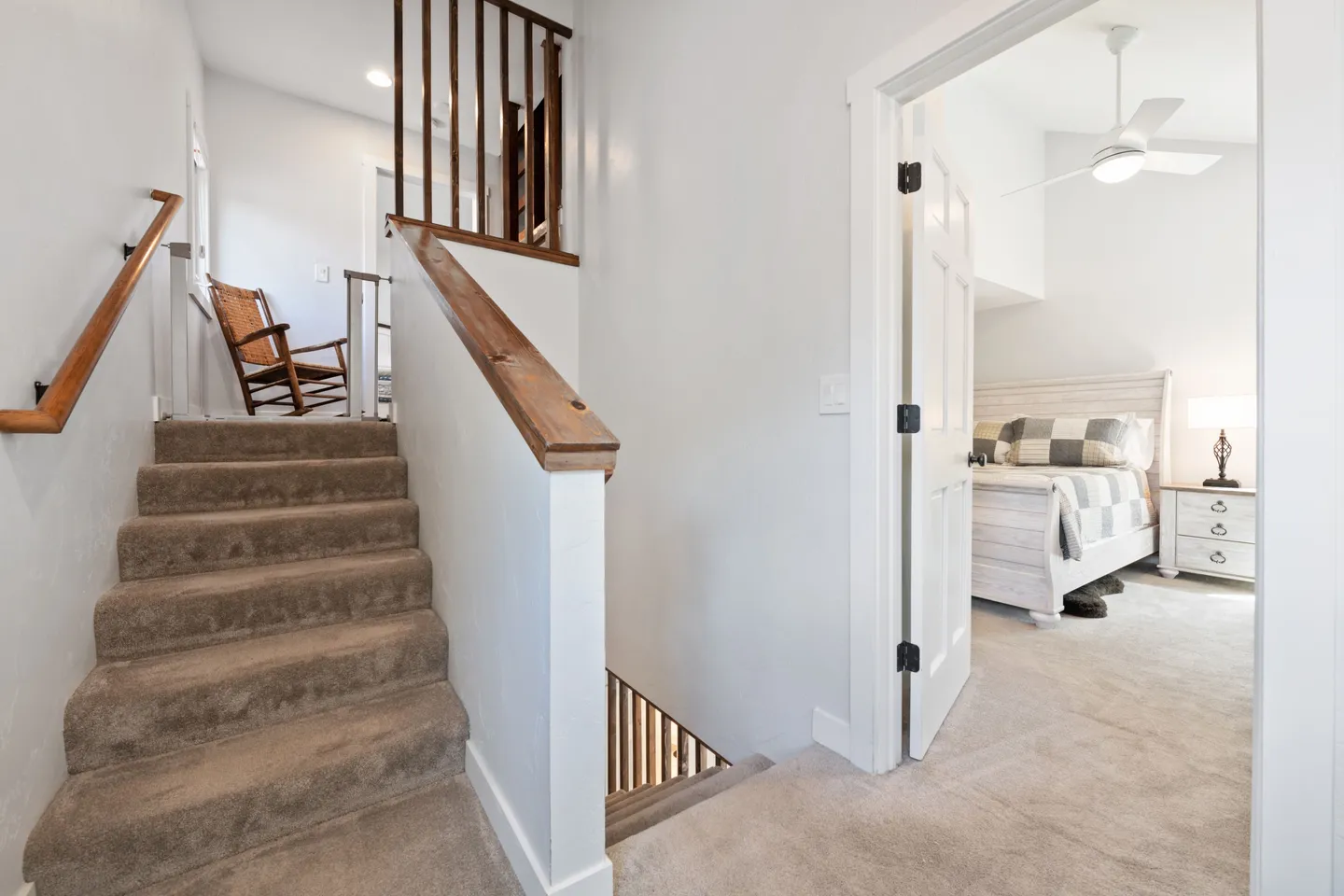 Carpeted landing with stairs leading up and down, adjacent to a bright bedroom with a bed and ceiling fan.