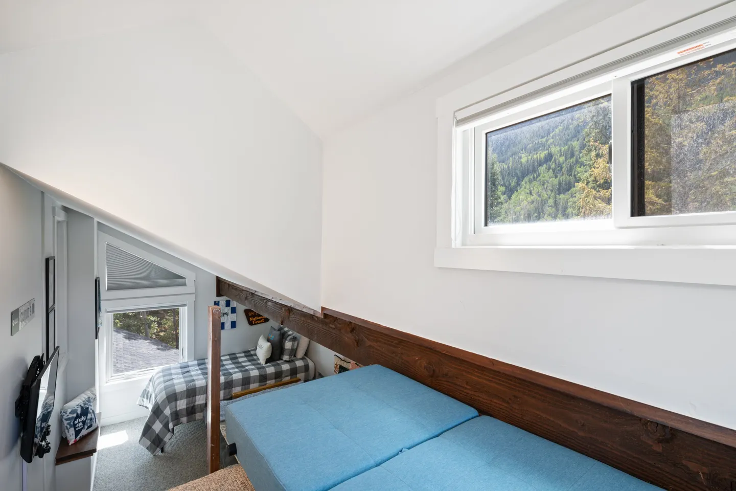 Elevated view of a compact loft sleeping space with blue mattresses, a wooden railing, and a view down into an adjacent bedroom area with a twin bed.