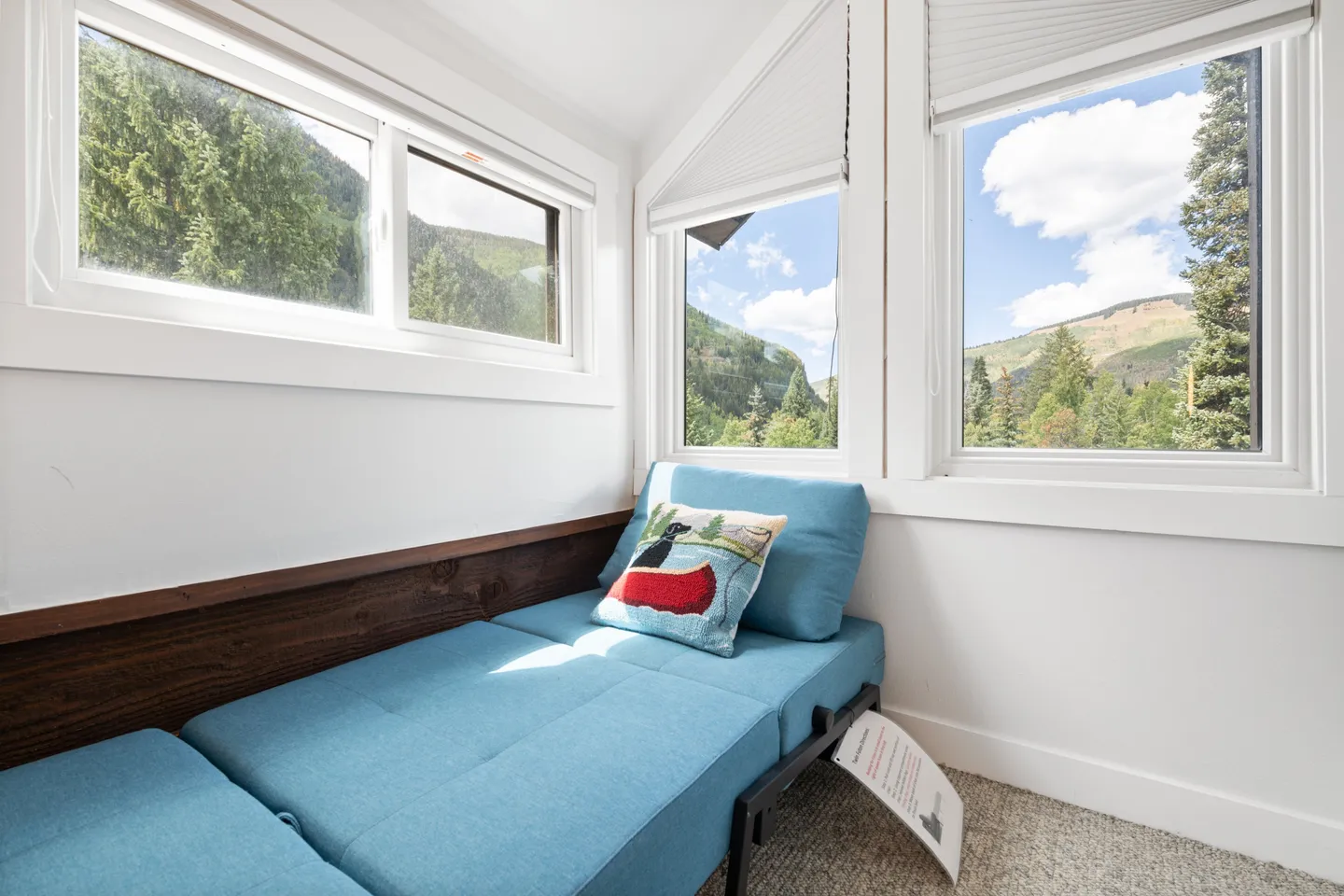 A cozy indoor window alcove featuring a low blue daybed cushioned with an embroidered pillow, surrounded by multiple windows showing forested hills and blue sky.