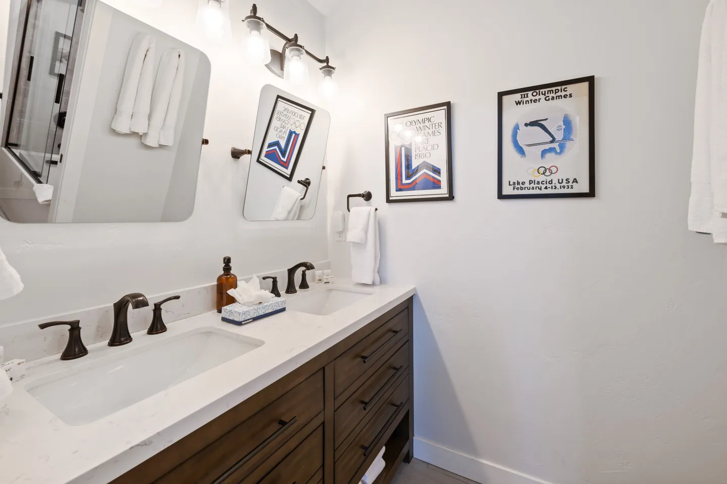 Bathroom vanity featuring a white countertop with two sinks, dark metal faucets, dark wood drawers, two mirrors, and Olympic posters on the adjacent wall.