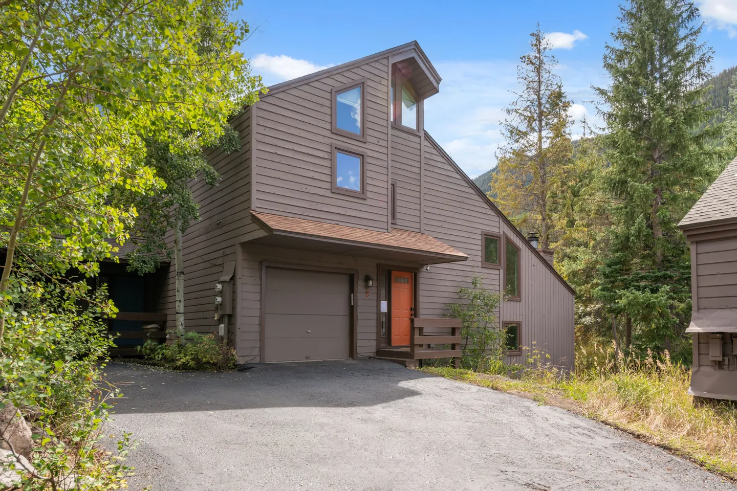 Exterior front of a brown multi-story home with an orange front door, single garage, and sloped roof against trees and blue sky.
