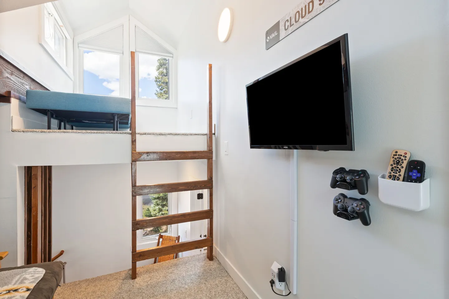 Carpeted loft area with a wooden ladder leading up to a raised sleeping nook, featuring a wall-mounted flat-screen TV with video game controllers and remotes.