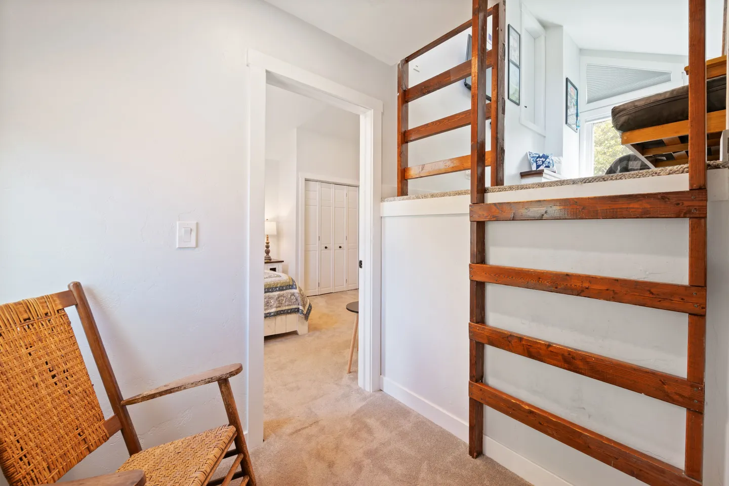 Carpeted hallway showing a rocking chair, an open doorway into a bedroom, and a wooden ladder leading up to a loft area.