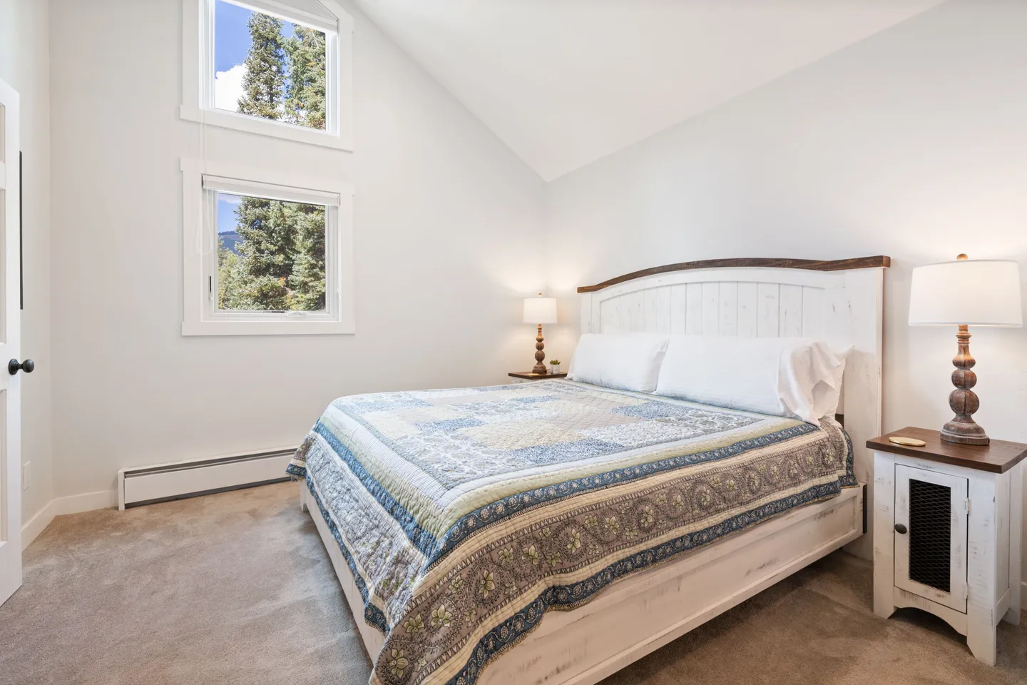 Bedroom with vaulted ceiling, carpeted floor, two stacked windows showing outdoor trees, and a wooden bed with nightstands.